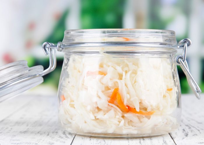 A clear jar of fresh sauerkraut with shredded carrot on a white wooden table