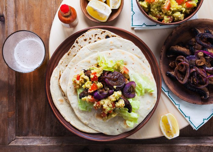 Plant-based meal of spiced mushroom fajitas set on a wooden table with other plates of fillings beside it