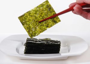 Woman lifting up a thin sheet of dried green seaweed from a pile of it on a white plate