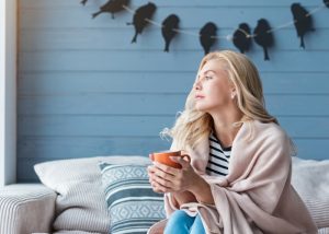 Woman sitting on her couch holding a cup of tea with a blanket draped over her shoulders