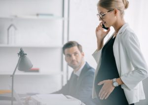 A pregnant female office worker on the phone in the office with a male colleague beside her