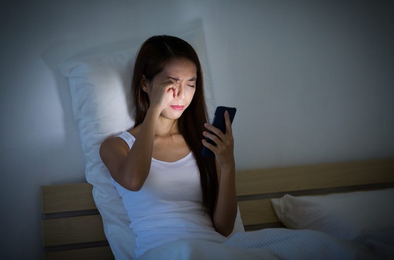 Woman rubbing her eyes while staring at her phone in bed at night and with glaring blue light