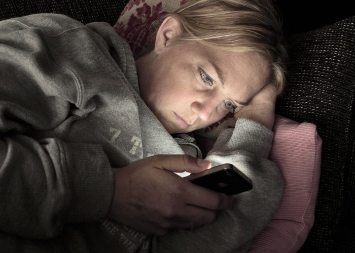 Woman in a hoodie jumper laying on her side on a couch in the dark and staring at her smartphone with glaring blue light