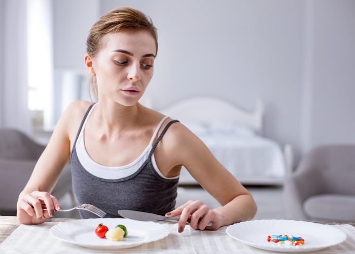 Slim woman holding a fork and knife over a plate with 3 pieces of food and looking at another plate filled with pills