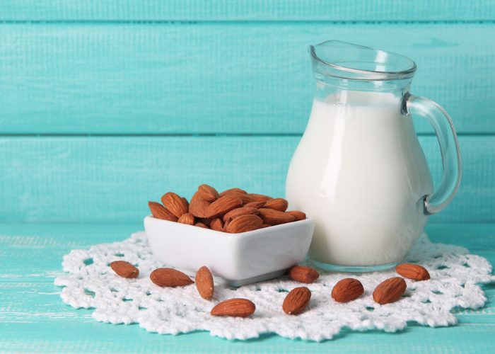 A jug of non-dairy almond milk with a square dish full of almonds next to it against a turquoise background