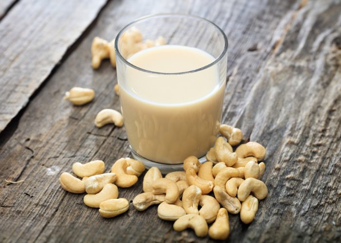 A glass of non-dairy cashew milk on a dark wooden table with cashews scattered around the glass