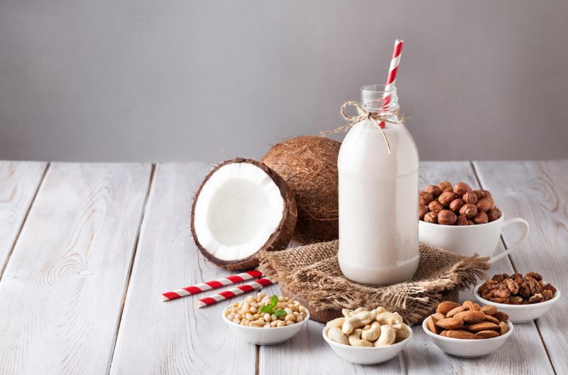 a bottle of non-dairy milk with a straw in it, surrounded by non-dairy milk sources like fresh coconuts, nuts, and grains