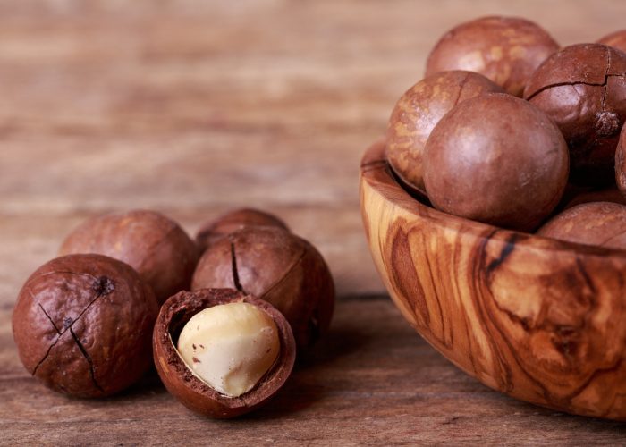 Close up of macadamia nuts on a wooden table, and half a wooden bowl full of macadamia nuts