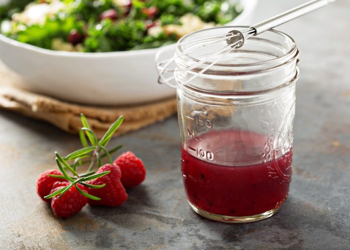 A jar of raspberry fruit vinegar with a whisky lying across the open mouth, on a table with a bowl of salad in the background and fresh raspberries next to the jar