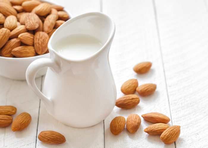 A jug of non-dairy almond milk, a bowl of almonds, and almonds scattered on the table around the jug