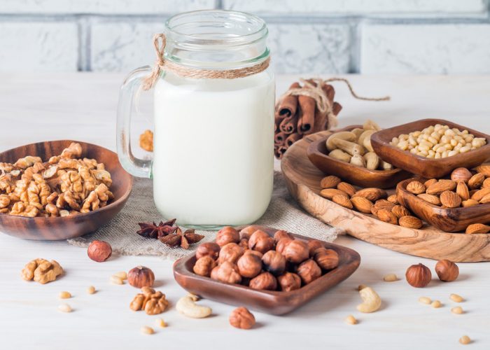 A mason jar filled with non-dairy milk and wooden dishes filled with nuts like walnuts, macadamias, cashews, and almonds around the jar on a white table