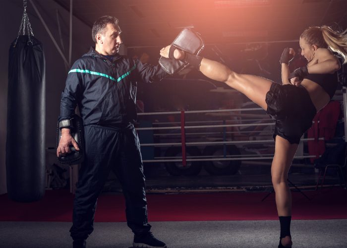 A fit blonde woman doing a high side kick with her kickboxing instructor holding a training pad