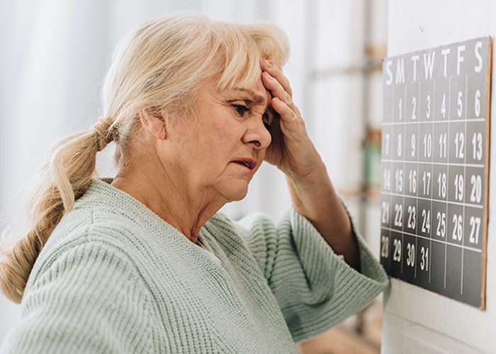 Menopausal old woman standing in front of a calendar holding her head as she struggles to remember