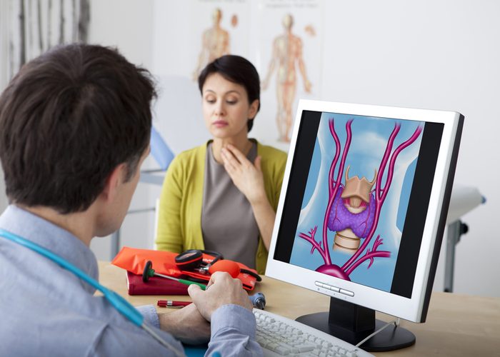 A doctor looking at a screen with an image of a woman's thyroid as she holds her neck in pain.