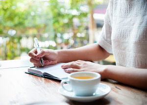 Woman journaling on a wooden table with cup of tea beside her