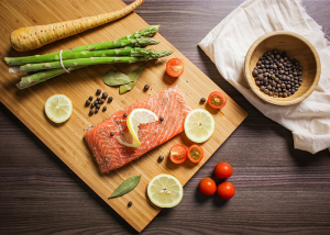 Top down view of pegan 365 diet (paleo-vegan) foods such as salmon, asparagus, carrot, lemons, and tomatoes on a wooden board being prepped for cooking