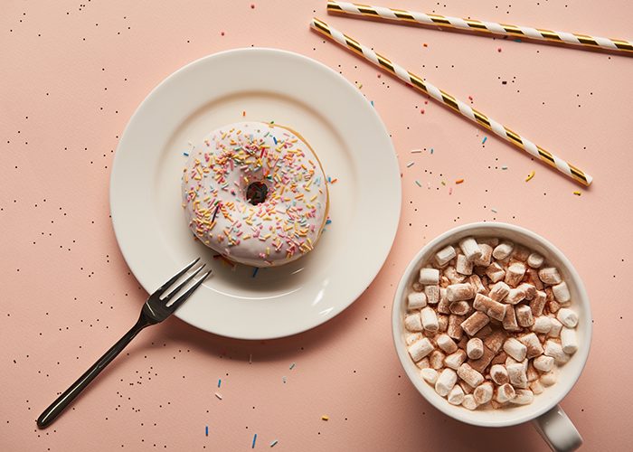 top down view of a donut with colourful sprinkles on a white plate with a fork on the side, two striped straws above it, and a mug of hot chocolate topped with cocoa powder and marshmallows in the bottom right corner