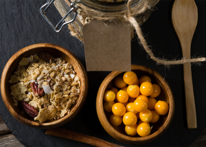 Top down view of a bowl of oats next to a bowl of golden berries and wooden spoon, with partial view of a jar.