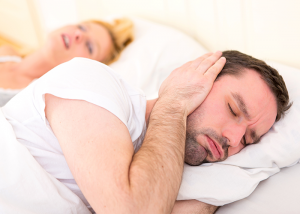 Couple in bed with woman snoring and man covering his ears.