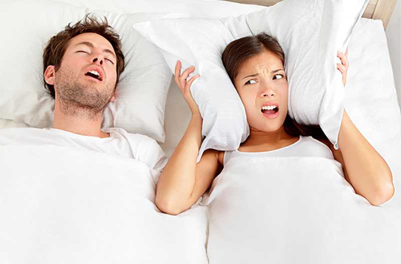 couple in bed with man snoring and woman looking annoyed and covering her ears with her pillow