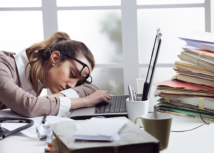 Woman with sleep disorder fast asleep at her office desk in front of her laptop and a stack of documents.