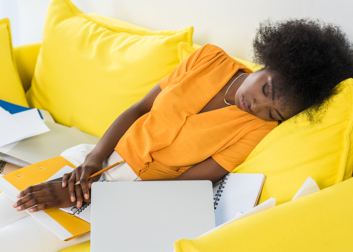 African American woman in yellow top asleep on a yellow couch with open notes, documents, and notepads on her lap.