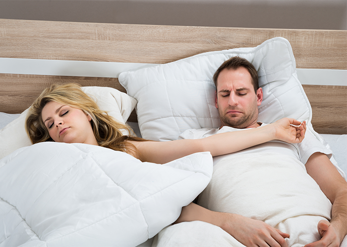 Couple in bed with man looking annoyed as his wife's arm is on him due to periodic limb movement disorder.