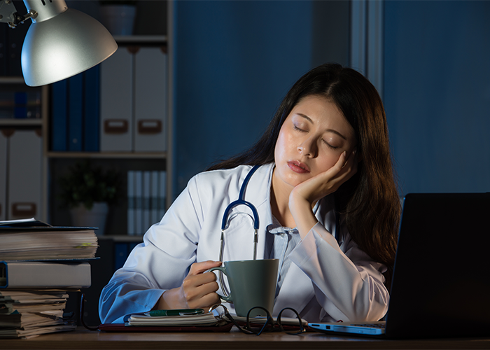 Doctor on shift with her head in her hands and a cup of coffee in the other falling asleep at her desk due to shift work sleep disorder.