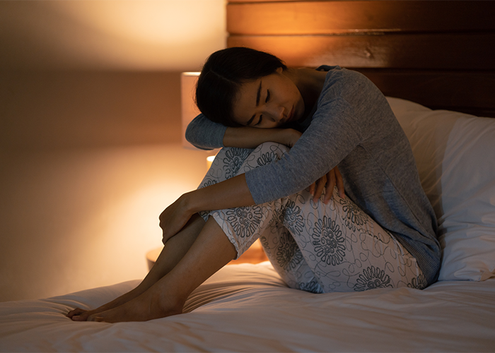 Woman with sleep disorder sitting up in bed at night with lamp on resting her head on her knees.