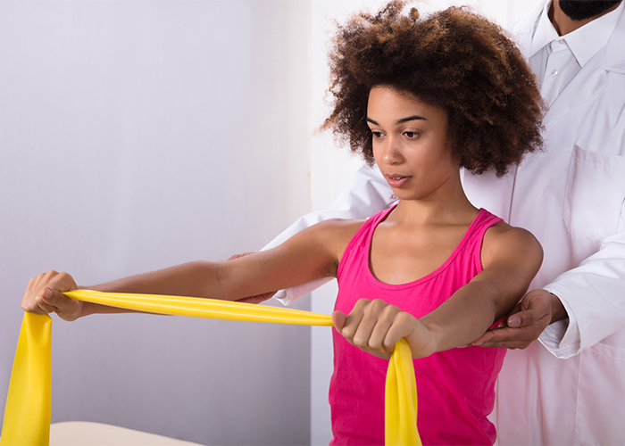 African American woman using resistance band to do shoulder exercises with the help of a physiotherapist