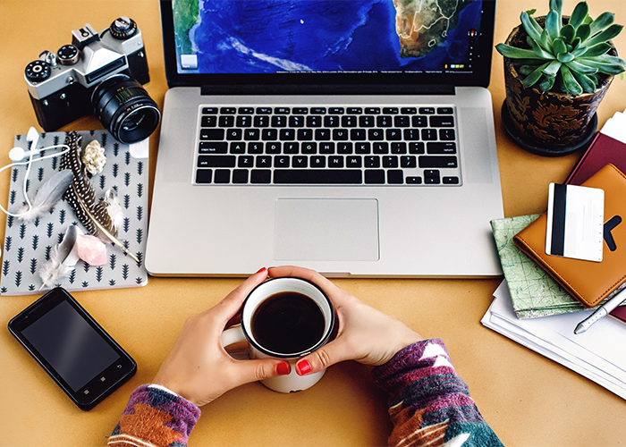 Woman's hands holding onto a cup of coffee with her laptop, camera, phone, a pile of notebooks, and a green plant in front of her