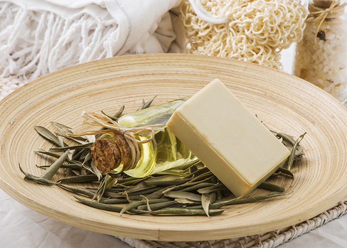 A goat milk soap bar on a bed of dried leaves, next to a bottle of essential oil, in a large dish