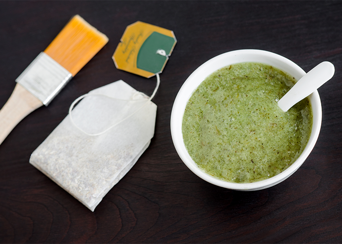 A bowl of green tea with a tea bag next to it as well as a facial brush