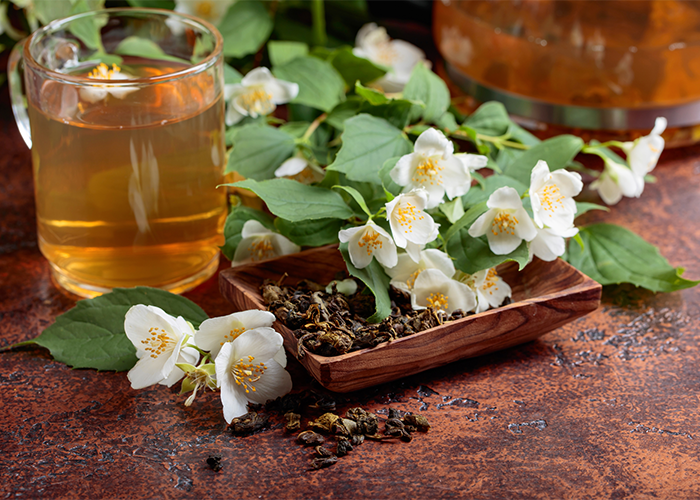 Cup of jasmine tea, a brown wooden dish with dried jasmine tea leaves, and fresh jasmine flowers