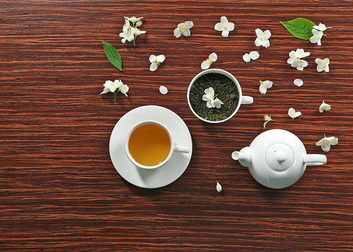 Top down view of a cup of jasmine green tea, a cup containing dried jasmine tea leaves, a white porcelain teapot, and scattered jasmine flowers