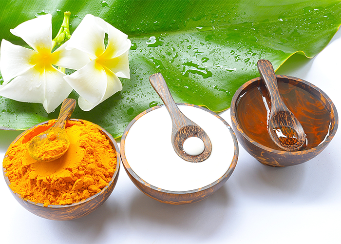 Turmeric powder, yogurt, and honey in wooden bowls with wooden spoons and a large green leaf and white flowers in the background