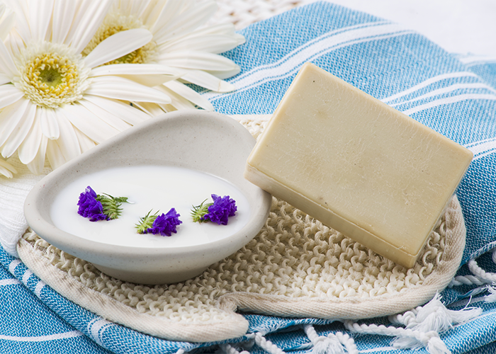 Goat milk soap bar next to a dish with white liquid soap and purple flowers, set on a blue cloth with white flowers behind