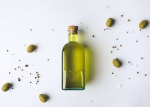 A bottle of olive oil lying flat on a white table, with olives surrounding it