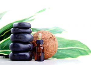 A bottle of coconut essential oil used for aromatherapy next to black stones and a whole coconut