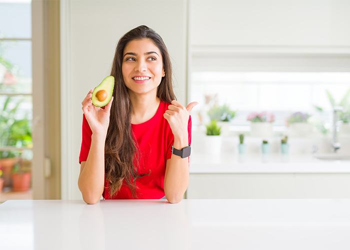 Woman in her kitchen holding up half an avocado and giving a thumbs up