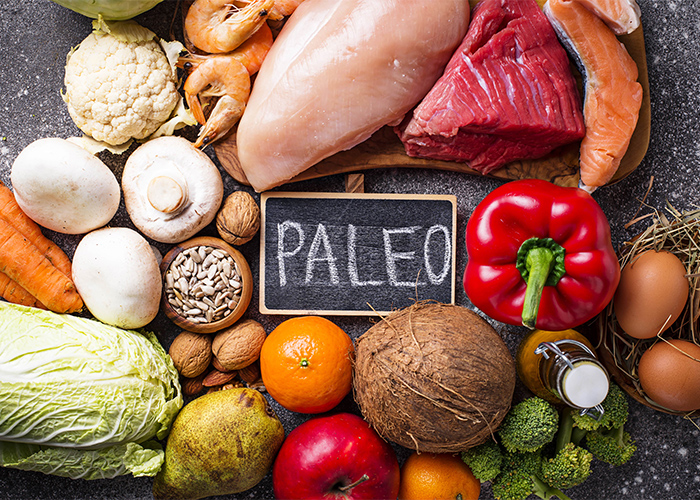 A top-down view of different foods on the Paleo diet including high protein low carbohydrate foods  like meat, poultry, nuts, vegetables, and fruit surrounding a small blackboard written with the word "PALEO"