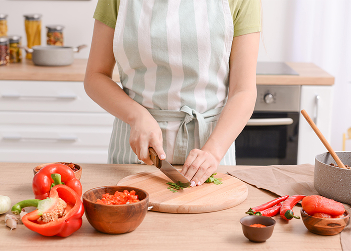 Woman in an apron in her kitchen preparing a Keto chili dish by cutting fresh herbs on a cutting board 